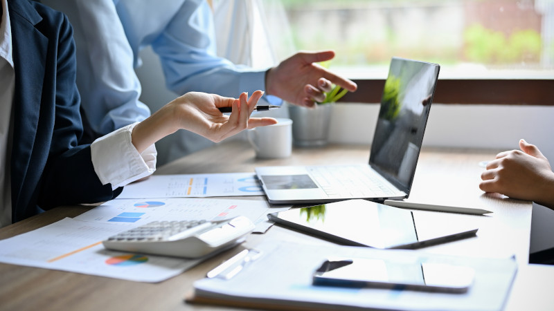 Stock image of people from shoulders down sat at table with laptop and business looking documents laid out.